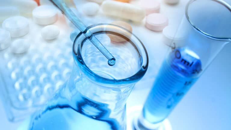 AstraZeneca pharmaceutical research laboratory depiction featuring a glass beaker with blue liquid, a dropper dispensing a drop, a graduated cylinder, and an assortment of pills and tablets in the background