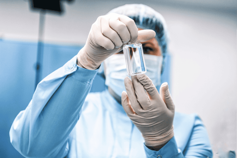 AstraZeneca pharmaceutical worker depiction wearing full cleanroom protective gear (blue lab coat, hairnet, face mask, and latex gloves) carefully inspecting a clear glass vial in a sterile manufacturing environment