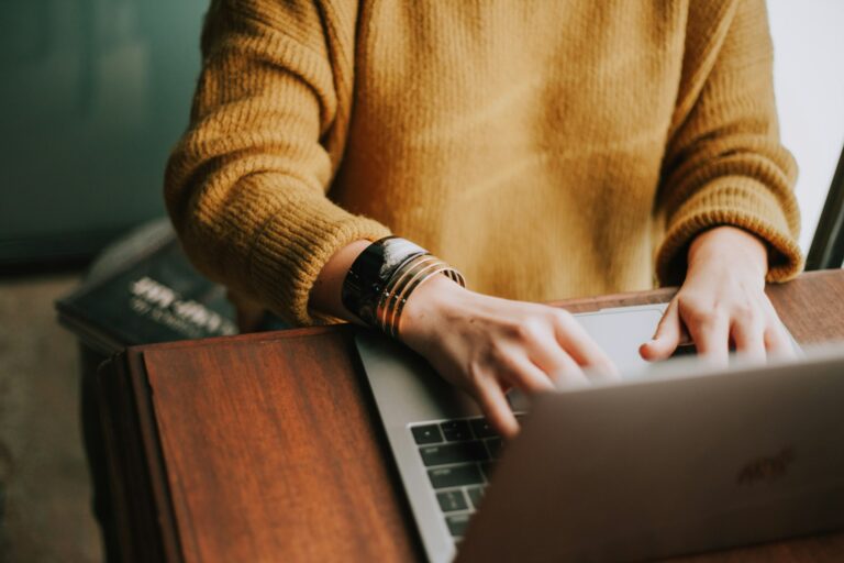 A person in a mustard yellow sweater is typing on a laptop at a wooden desk, possibly working on SAP or preparing for Questionmark assessments. Only their arms and hands are visible, and they are wearing multiple bracelets.