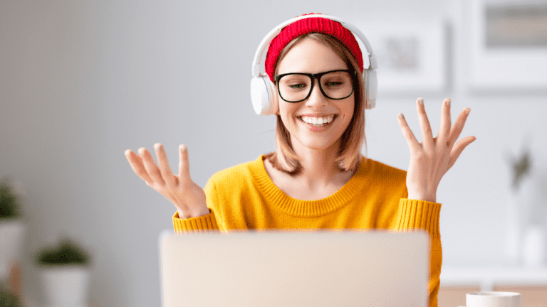 A smiling person in glasses, a red beanie, and headphones gestures with both hands while looking at a laptop, perhaps engaging with innovative employee assessment tools. The background is softly blurred, suggesting a cozy indoor setting.