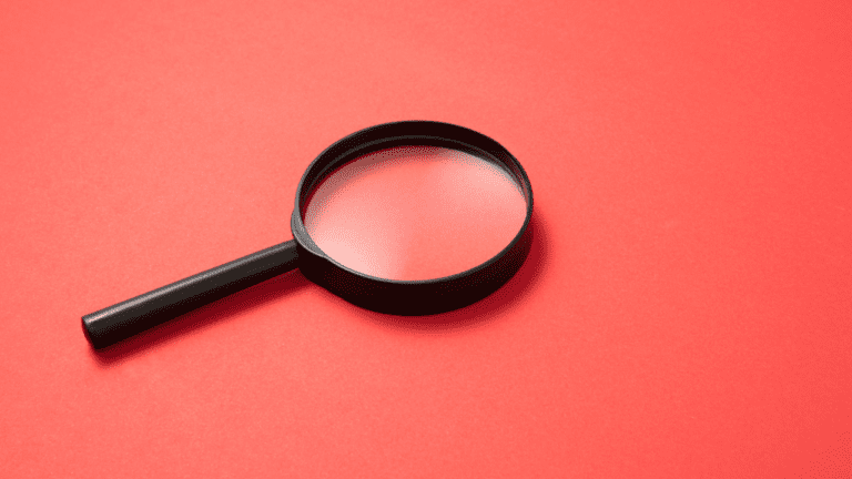 A black magnifying glass lies on a flat red surface, angled diagonally with the handle pointing left, evoking the careful scrutiny found in observational assessments.