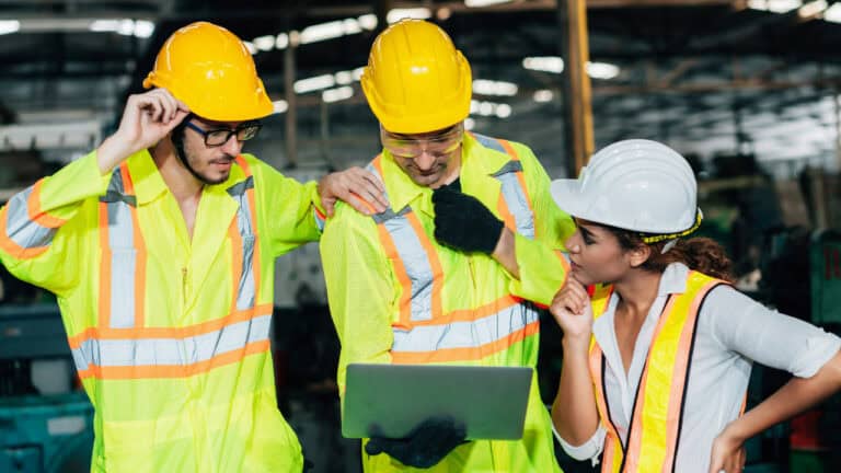 Three people in hard hats and safety vests are looking at a laptop