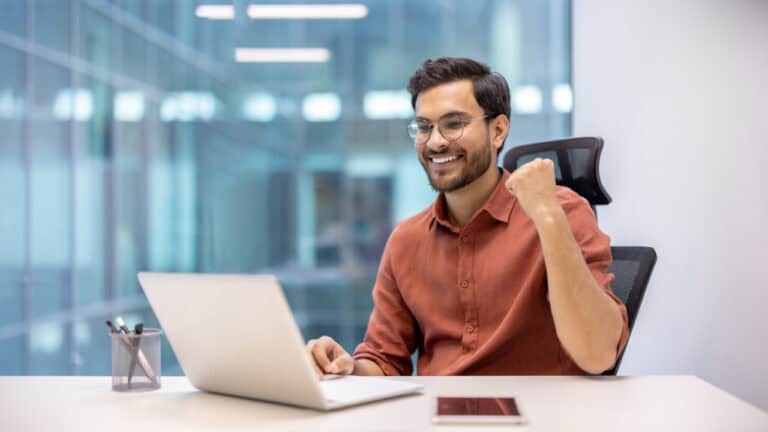 A man wearing glasses and an orange shirt sits at a desk, smiling and making a fist pump while looking at his laptop in a modern office, celebrating his successful scale certification.