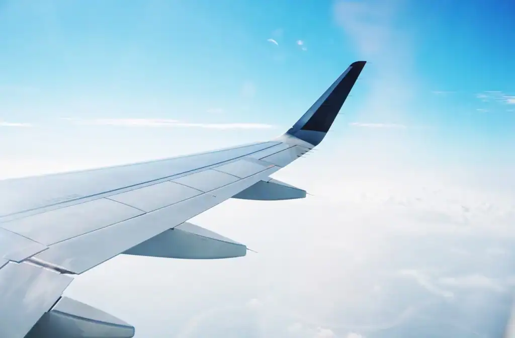 An airplane wing flying over a vast blue sky with clouds underneath.