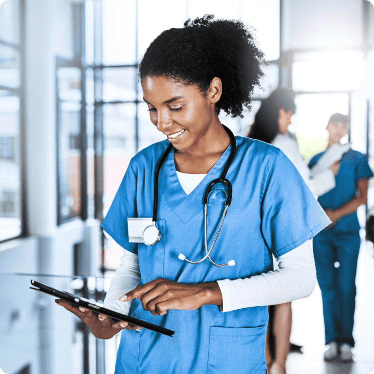A healthcare professional in blue scrubs, wearing a stethoscope, smiles while using a tablet in a bright medical facility that upholds strict compliance and regulation. Other people in medical attire are visible in the background.