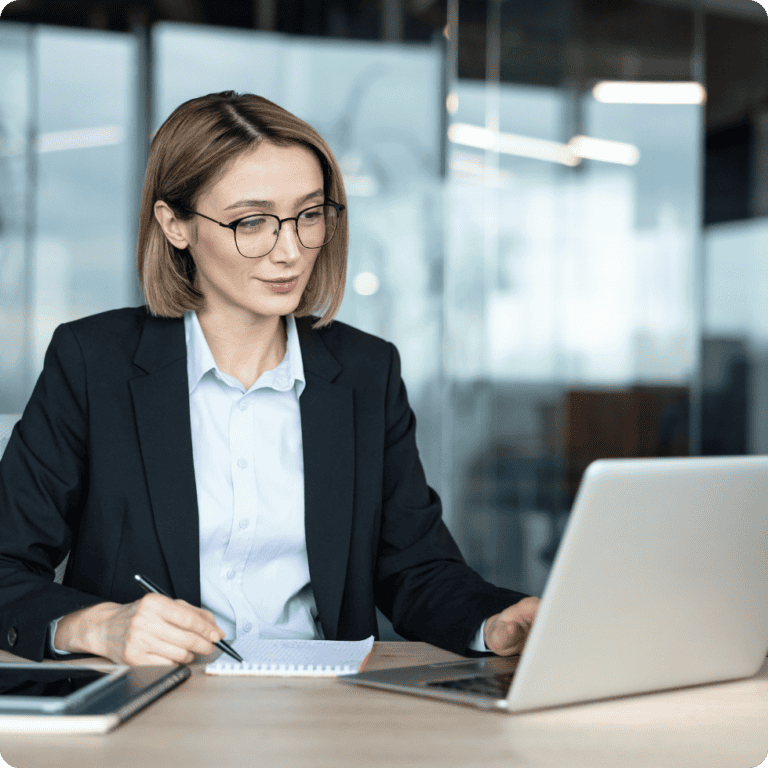 A woman in business attire sits at a desk with a laptop, taking notes in a notebook. She wears glasses and is focused on her work in a modern office setting, reviewing employee assessment tools to enhance workplace productivity.